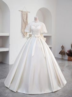 Laced with the nostalgia of a bygone era, this wedding dress boasts a high-neck lace bodice that radiates elegance and poise. Its delicate sleeves brush the edges of the shoulders, a soft prelude to the voluminous satin skirt that cascades into a regal train. The simplicity of the silhouette is its grandeur, tied together by a satin sash at the waist, making this gown a timeless choice for a bride's unforgettable day. Wedding Dresses With Neck Collar, Unique Ball Gowns, Silver Decorations, Satin Long Prom Dress, Formal Wedding Dress, Lace Top White, डिजाइनर कपड़े, Top In Pizzo, Satin Formal Dress