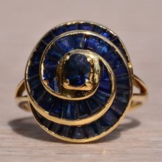 The Willare: Vintage Spiral Sapphire And Yellow Gold Cocktail Ring. Admire This Impressive Cocktail Ring Set With 2.50 Total Carats Of Blue Sapphires Set In A Fibonacci Spiral Design. At The Heart Of The Spiral Is An Oval Deep Blue Sapphire Followed By Tapered Baguettes Throughout The Rest Of The Ring. The Ring Is Crafted In 14 Karat Yellow Gold And Is Currently A Finger Size 8.5 And Is Able To Be Resized For An Additional Fee Upon Request. Yellow Gold Cocktail Ring, Fibonacci Spiral, The Spiral, Gold Cocktail Ring, Gold Cocktail, Spiral Design, Blue Sapphire Rings, Cocktail Ring, Cocktail Rings