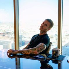 a man with tattoos sitting at a desk in front of a window looking out on the city