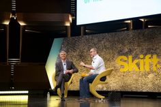 two men sitting on chairs in front of a screen with skift's logo