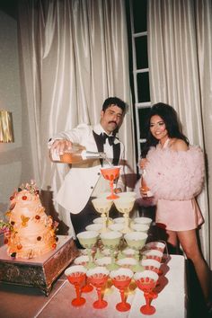 a man and woman standing in front of a table filled with cakes, drinks and desserts