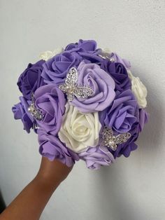 a bridal bouquet with purple and white flowers is held up against a wall by someone's hand