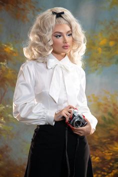 A blond pinup model posing against a yellow and blue painted background while wearing the Angela Blouse in white and black trousers with an old camera on her hand. Classic White Satin Blouse, Fall White Blouse With Tie Sleeves, White Tie Sleeve Blouse For Fall, White Tie Sleeves Blouse For Fall, White Blouse With Tie Sleeves For Fall, Feminine Fitted Blouse With Bow, White Bow Blouse For Work, Fitted Feminine Blouse With Bow, White Bow Blouse For Workwear