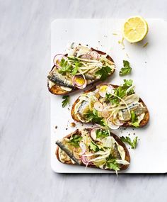 three pieces of bread with fish and vegetables on it next to a lemon wedges