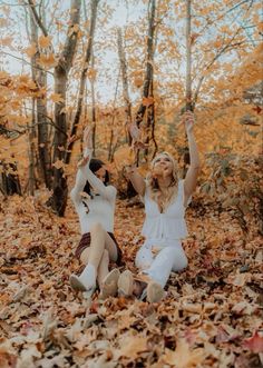 two people sitting on the ground surrounded by leaves and trees with their arms in the air