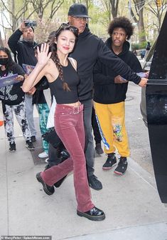 a woman in red pants and black top walking down the street with other people behind her