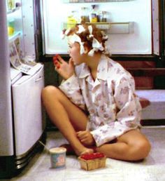 a woman is sitting on the floor in front of an open refrigerator and eating something