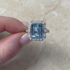 This gemstone ring features a 3.26 carat emerald-cut aquamarine, bezel-set and accented by an old European cut diamond halo. Baguette cut diamonds are set flanking the center stone. A row of old European cut diamonds adorns each shoulder. The total diamond weight of this ring is approximately 0.62 carats. This ring is handcrafted in platinum, decorated with fine milgrain and an openwork under-gallery.
The measurements of the aquamarine are approximately 10.06mm x 8.07mm x 5.43mm. The approximate Baguette Cut Topaz Ring With Diamond Halo Setting, Baguette Cut Topaz Ring With Diamond Halo, Gia Certified Emerald Cut Topaz Ring In Platinum, Gia Certified Emerald Cut Platinum Topaz Ring, Fine Jewelry Emerald Cut Topaz Ring Gia Certified, Heirloom Diamond Topaz Ring With Emerald Cut, Dazzling Emerald Cut Diamond Topaz Ring, Heirloom Diamond Topaz Ring, Emerald Cut, Emerald-cut Topaz Ring With Halo Setting