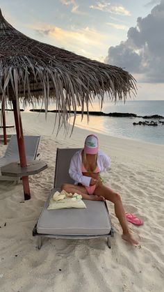 a woman sitting on top of a beach chair under a thatched umbrella next to the ocean