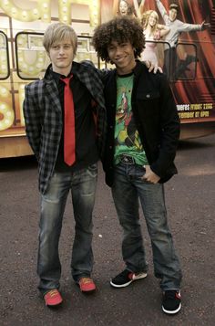 two young men standing next to each other in front of a movie poster and bus