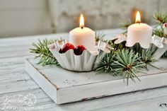 three lit candles sitting on top of a white tray filled with pine cones and berries