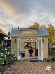 a small yellow shed sitting in the middle of a gravel lot with flowers growing around it