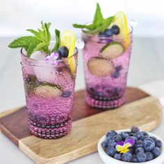 two glasses filled with blueberries and lemonade on a wooden tray next to a bowl of fruit