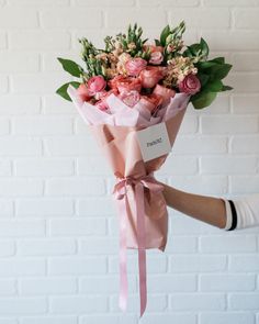 a person holding a bouquet of flowers in front of a white brick wall with pink ribbon