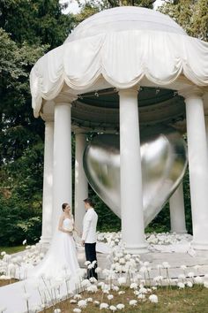 a bride and groom standing in front of a large heart shaped structure with white flowers