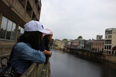 two people are sitting on a ledge overlooking a river and buildings in the distance, with one person wearing a white hat