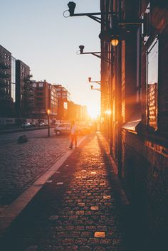 the sun is setting on an empty street with people walking down it and buildings in the background