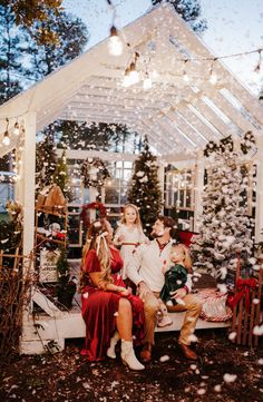 three people sitting on a bench in front of a christmas tree with snow falling all around them