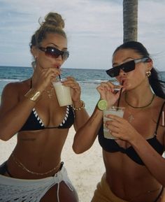 two beautiful women standing next to each other on a beach drinking drinks from cups in front of the ocean