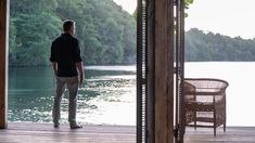 a man standing on a wooden deck looking out at the water from inside an open door