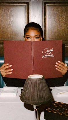 a woman sitting at a table with a book in front of her face while holding it up