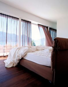 an unmade bed sitting in front of two large windows with sheer drapes on them