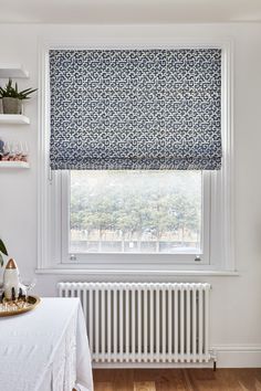 a room with a table, radiator and window in the corner that has a blue patterned roman blind on it