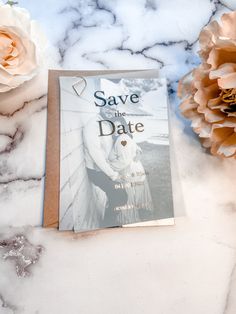 a save the date card sitting on top of a marble table next to two flowers