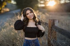 a beautiful young woman standing next to a wooden fence in a field with tall grass