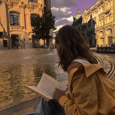 a person sitting on a bench reading a book in front of a fountain with water spouting from it