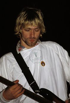 a man with long blonde hair holding a guitar in his right hand and wearing a white shirt