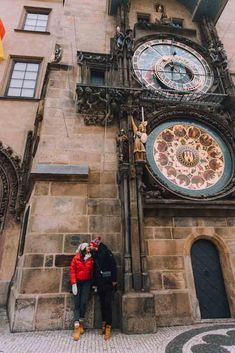 two people standing next to each other in front of a building with clocks on it