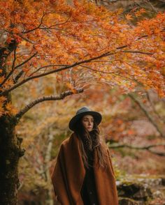 a woman wearing a brown coat and hat standing in front of a tree with orange leaves