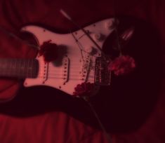 a white electric guitar sitting on top of a red bed sheet with flowers in the foreground