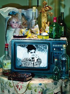 an old tv sitting on top of a table covered in bottles and other items next to a statue