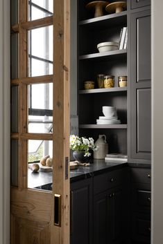 an open door leading to a kitchen with black cabinets and white dishes on the shelves