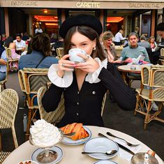 a woman sitting at a table drinking from a cup with food on the plate in front of her