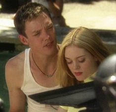 a young man and woman sitting next to each other looking at something on the ground