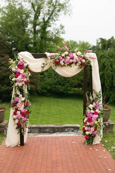 a wedding arch decorated with pink flowers and greenery on the side of a brick walkway