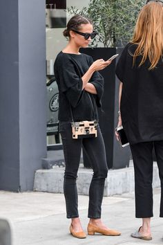 two women are standing on the sidewalk looking at their cell phones and one is holding a purse