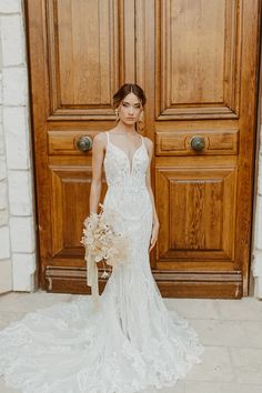 a woman standing in front of a wooden door wearing a wedding dress and holding a bouquet