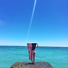 an american flag draped over a person standing on a rock in the ocean with blue sky