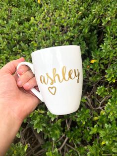 a hand holding a white coffee cup with gold foil lettering on it, in front of some bushes