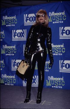 the blond haired woman is dressed in shiny black clothing and holding a handbag while posing for