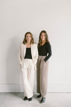 two women standing next to each other in front of a white wall wearing pants and jackets