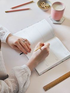 a woman sitting at a table writing in a notebook with pencils and paper on it