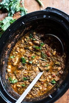 a crock pot filled with beans and greens on top of a wooden table next to a spoon