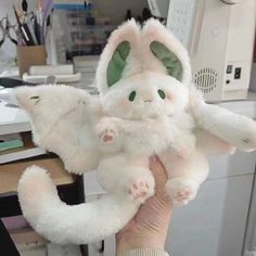 a hand holding a white stuffed animal with green eyes and tail, in front of a computer desk