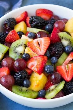 a white bowl filled with fruit on top of a wooden table next to grapes, kiwis and strawberries