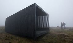 two people standing next to a black structure on top of a grass covered hill in the fog